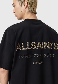 Zwarte oversized t-shirt met gouden luipaardprint tekst "ALLSAINTS" en extra tekens. Losse pasvorm met korte mouwen en ronde hals.