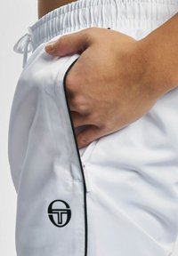 Weiße Sportshorts mit strukturierter Oberfläche, ausgestattet mit einem schwarzen Reißverschluss und Besatz sowie einem Logo-Patch an der Seitentasche.