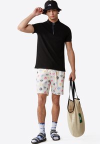 Czarna koszulka polo z niebieskimi akcentami, białe szorty w kolorowe wzory, czarny kapelusz typu bucket, niebieskie skarpetki tie-dye oraz beżowa torba na ramię z czarnymi uchwytami.