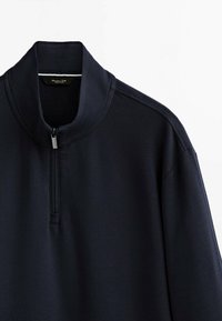 Pull noir à manches longues avec un col montant et une courte fermeture éclair sur le devant, en tissu lisse, suspendu sur un fond blanc.