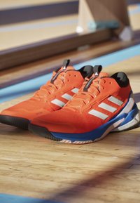 Orange athletic shoes with a textured upper, white stripes, blue sole accents, and black heel. Features a supportive design for active wear.