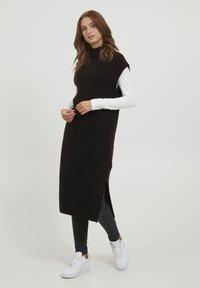 Robe sans manches en tricot noir avec un col montant, fentes latérales et texture côtelée, portée par-dessus une chemise blanche à manches longues et un pantalon noir.