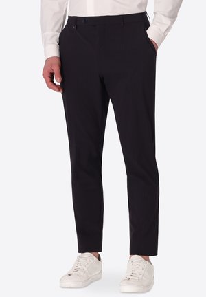 Uomo che indossa pantaloni neri slim-fit con tasche laterali, camicia bianca e sneakers bianche, in piedi con una mano in tasca.