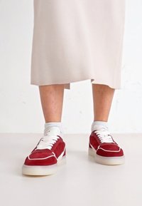 Sneakers in suede rosso con accenti e lacci bianchi. Presentano una linguetta imbottita e una suola traslucida. Indossati con calzini bianchi e una gonna neutra.
