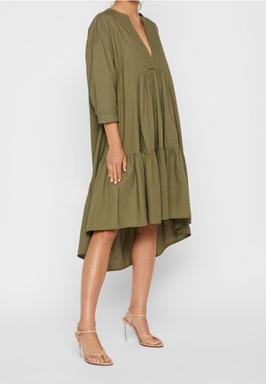 Robe en coton vert olive avec un décolleté en V, des manches trois-quarts et un ourlet en couches. Associée à des talons nus à lanières.