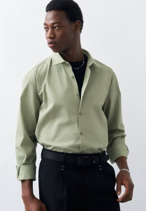 Jeune homme regardant vers la gauche, portant une chemise boutonnée vert clair par-dessus un haut noir, un pantalon noir avec une ceinture, ainsi qu'une chaîne en argent et un bracelet.