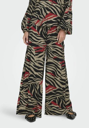 Pantalons beige amples avec des rayures zèbres noires et rouges, présentant une coupe large, associés à des ballerines noires cloutées.