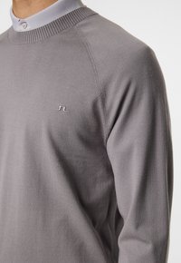 Grå stickad tröja med rund halsringning och ribbad krage. Har en liten, ton-i-ton logotyp på vänster bröst och en mjuk textur.