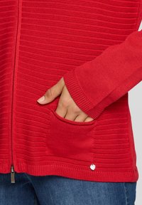 Roter Cardigan mit Rippenmuster, Reißverschluss, Seitentaschen und kleinem Anhänger. Verfügt über eine strukturierte Webart und figurbetonte Ärmel.