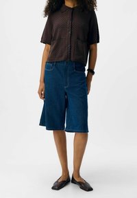 Chemise marron en maille à manches courtes avec deux poches avant. Short Bermuda en denim bleu à coupe droite, doté de passants de ceinture et de coutures visibles.