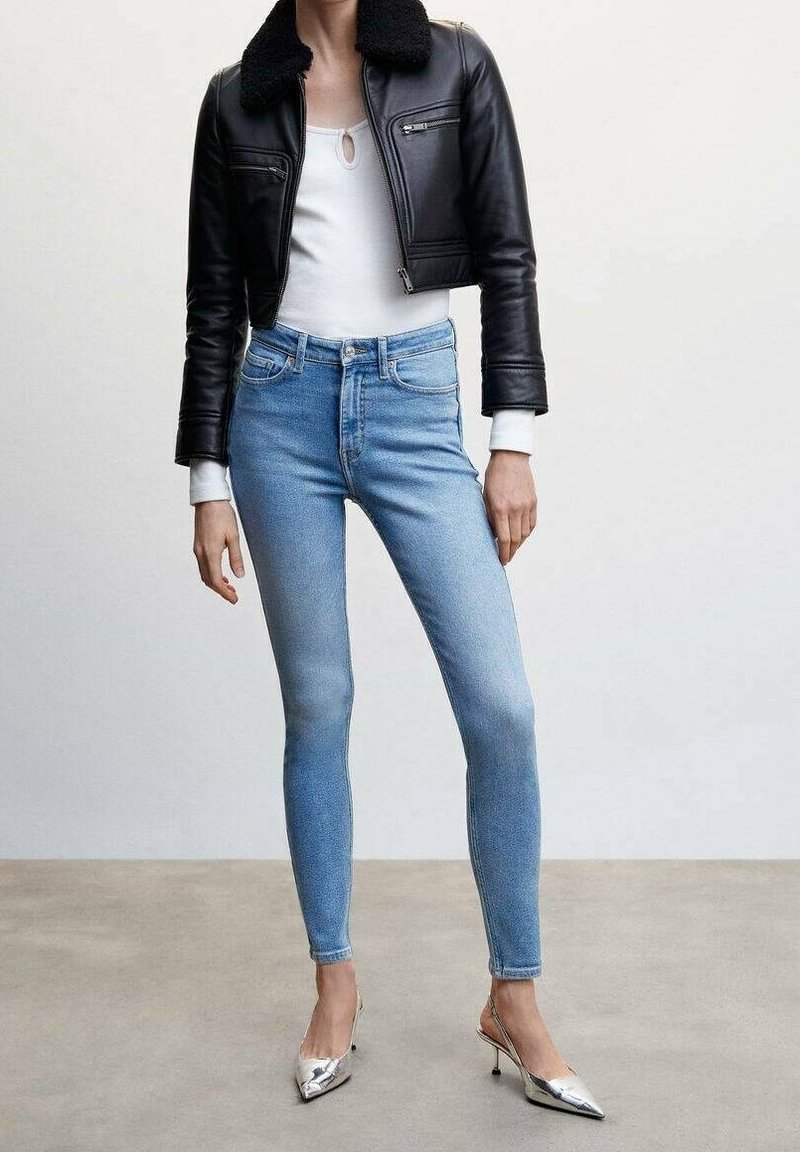 Veste courte en cuir noir avec un col en peau lainée, haut blanc, jean skinny bleu clair et talons métalliques à bout pointu.