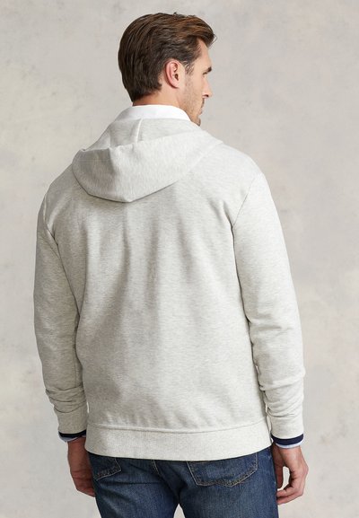 Grå huvtröja med huva, ribbade muddar och fåll. Mjukt, texturerat tyg. Buren över en vit skjorta med krage och matchad med blå jeans.