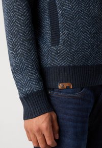 Pulover tricotat de culoare bleu marin cu model în zigzag, manșete cu nervuri și un buzunar lateral. Etichetă din piele maro cu inscripția "EST. 1993" pe blugi.