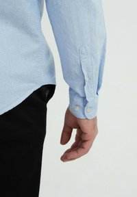 Chemise bleu clair à manches longues, avec un poignet à un bouton et un ourlet arrondi, associée à un pantalon noir.