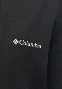 Μαύρο ύφασμα με λευκό λογότυπο της μάρκας Columbia τυπωμένο στο στήθος ενός ενδύματος.