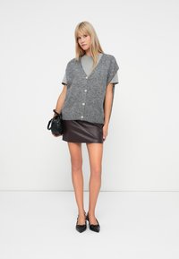 Šedý pletený vestový sveter s krátkymi rukávmi a prednými gombíkmi, vrstvený cez svetlo šedú košeľu a tmavo hnedú mini sukňu z umelého kože. Čierna kabelka a topánky.