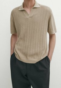 Polo en maille beige à manches courtes avec motif côtelé, porté avec un pantalon noir, mains dans les poches, sur une personne devant un fond clair uni.