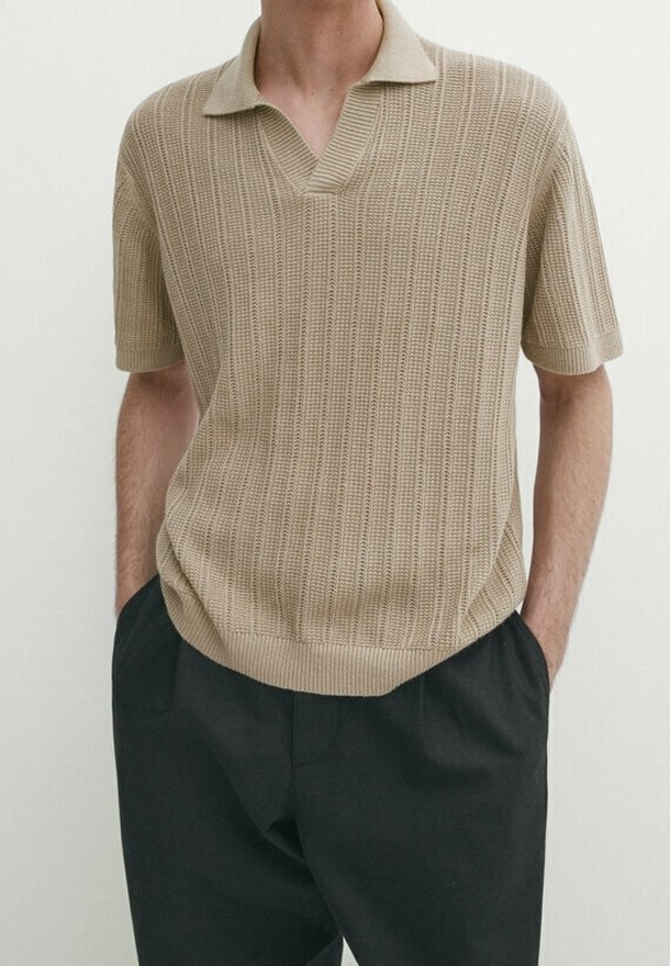 Polo en maille beige à manches courtes avec motif côtelé, porté avec un pantalon noir, mains dans les poches, sur une personne devant un fond clair uni.
