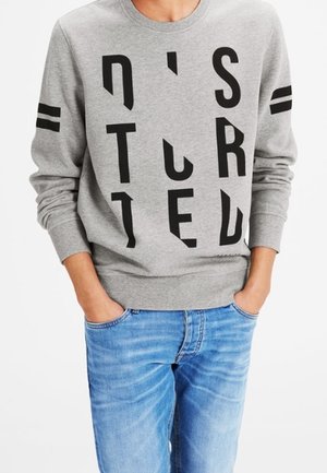 Person iført en grå sweatshirt med sorte abstrakte lodrette bogstaver og to sorte striber på hver ærme, kombineret med blå jeans.