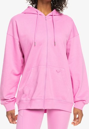 Femme portant un sweat à capuche zippé rose clair avec cordons et poches avant, assorti à des leggings roses assortis.