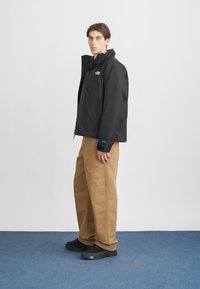 Μαύρο αδιάβροχο μπουφάν με ψηλό γιακά, συνδυασμένο με καφέ παντελόνια φαρδιάς γραμμής και μαύρα αθλητικά παπούτσια με λογότυπα και υφή.