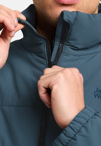 Giacca a vento teal con collo alto, chiusura con zip e polsini elastici. Presenta un logo sul petto e un tessuto liscio e testurizzato.