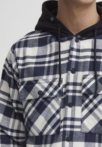 Camicia in flanella a quadri blu navy e bianca con cappuccio nero, dotata di due tasche frontali, bottoni a pressione e un design del tessuto testurizzato.