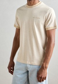 Beige bomuld t-shirt med rund hals, korte ærmer og kontrasterende brun kant. Har diskret branding på brystet.