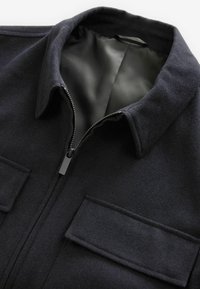 Giacca di lana blu navy con colletto semplice, chiusura con zip e due tasche frontali. Interno foderato in tessuto nero liscio.