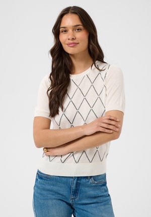 Femme aux longs cheveux bruns, debout les bras croisés, porte un pull blanc à manches courtes à motif losange noir et un jean bleu.