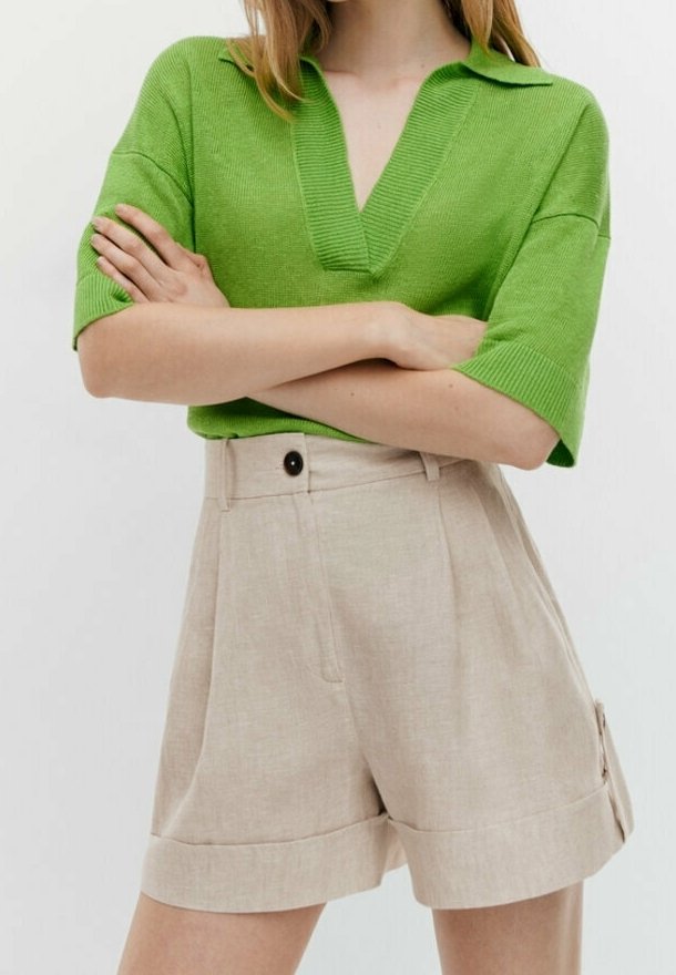 Personne portant un pull vert vif à manches courtes et col en V, ainsi qu'un short beige taille haute avec un bouton et des ourlets revers, bras croisés.