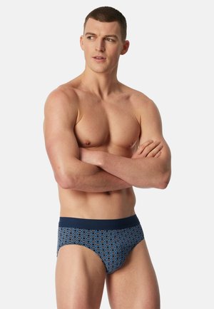 Hombre musculoso con cabello corto, vistiendo calzoncillos azules con estampado, de pie con los brazos cruzados, mirando hacia un lado sobre un fondo liso.