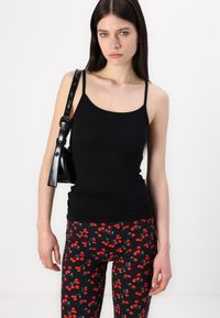 Svart tajt linne med tunna remmar tillsammans med svarta leggings med ett mönster av röda körsbär. Bakgrunden är helt vit.