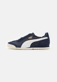 Zapatillas azul marino con una franja blanca, acentos de ante en la parte trasera y una suela de goma texturizada. Detalle del logo en dorado en el costado.