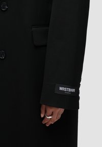 Cappotto nero su misura con chiusure a bottone, cuciture dettagliate e un'etichetta che recita "WRSTBHR STUDIO" sul polsino. Texture liscia.