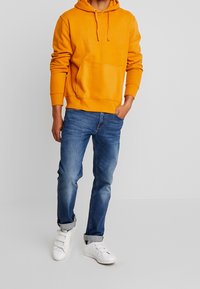 Hombre con sudadera naranja, jeans azules doblados y zapatillas de velcro blancas, de pie con una mano en el bolsillo sobre un fondo liso.