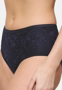 Prim-plan cu o persoană care poartă chiloți de damă cu talie înaltă, din dantelă florală în nuanțe de albastru închis, având un tiv neted.