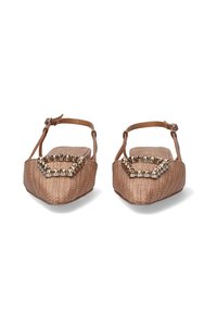Zapatos planos de punta abierta en material trenzado color marrón claro. Cuenta con un detalle decorativo de tachuelas metálicas en la parte frontal. Incluye una correa para el tobillo.