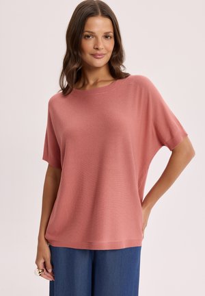 Kobieta ubrana w luźny, różowawy sweter z krótkim rękawem i niebieskie spodnie, stojąca z jedną ręką na biodrze na jednolitym tle.