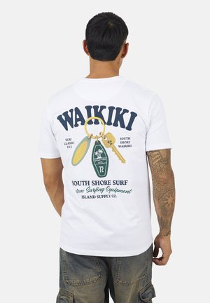 Uomo che indossa una maglietta bianca con la scritta "Waikiki" e un grafico di un portachiavi, insieme a del testo riguardante South Shore Surf e l'attrezzatura per il surf sul retro.