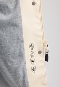 Gray fabric interior with textured surface; outer layer in cream featuring round snaps and five icons for windproof, heated seams, waterproof, and two-way zip.