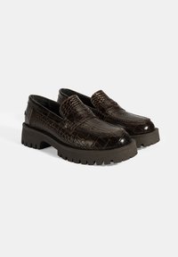Bruine loafers met krokodillenpatroon en een glanzende afwerking, voorzien van een dikke zwarte rubberen zool en textuurelementen op de bovenkant.