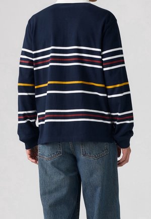 Person iført en navyblå sweater med hvide, røde og gule vandrette striber og blå denimjeans, vist bagfra.