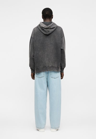 Férfi kopott fekete kapucnis pulóverben és laza világoskék farmernadrágban, háttal áll egy egyszínű fehér háttér előtt.