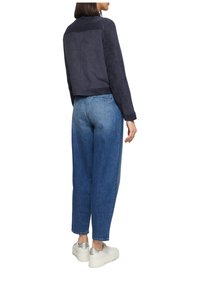 Vrouw in een donkerblauwe jas, met blauwe high-waisted jeans en witte platform sneakers, staand en met de rug naar de camera tegen een witte achtergrond.