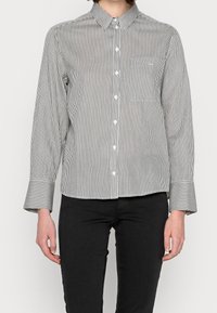 Chemise boutonnée à rayures noires et blanches avec col, poche poitrine unique et poignets évasés. Le tissu semble lisse et léger.
