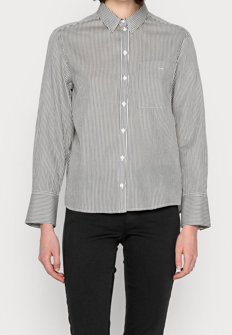 Chemise boutonnée à rayures noires et blanches avec col, poche poitrine unique et poignets évasés. Le tissu semble lisse et léger.
