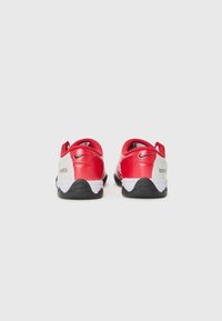 Chaussures de sport rouges avec une tige en synthétique lisse, des accents blancs, un talon texturé et une semelle en caoutchouc noir avec un détail de logo circulaire.