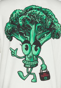 White t-shirt featuring a cartoon broccoli character in green, holding a black bag with red text "des légumes". Graphic design is bold and playful.