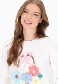 Fehér pulóver színes, kötött virágapplikációkkal és rózsaszín szalagdíszítéssel. A modell hosszú, hullámos barna hajjal rendelkezik, és virágos fülbevalót visel.
